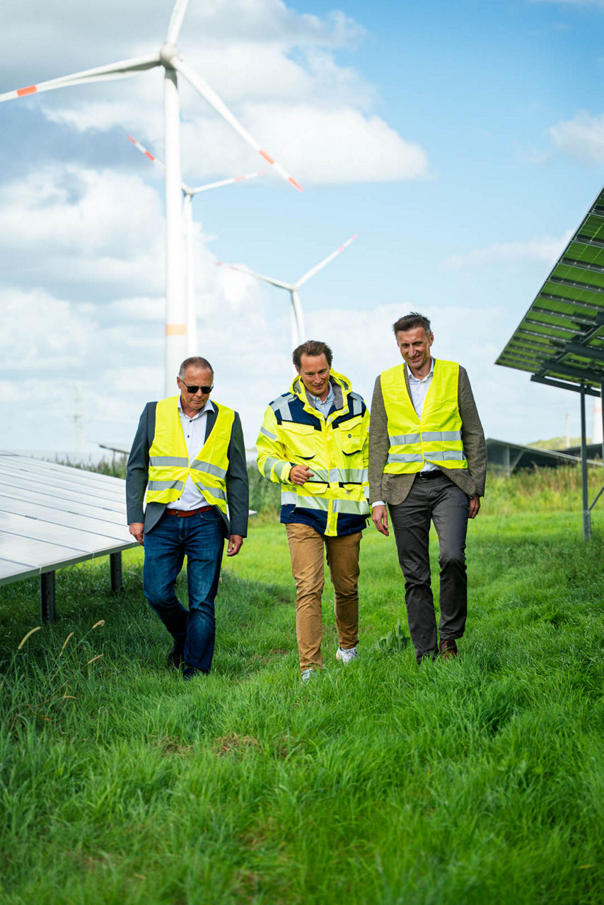 Drei Personen im Solarpark mit Windrädern im Hintergrund