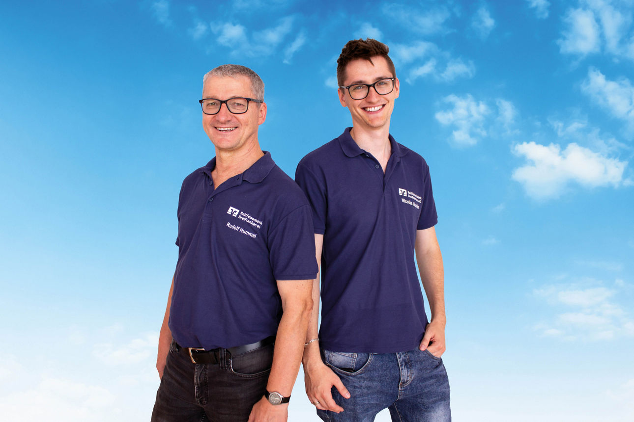 Die Kollegen Rudi Hummel und Nicolas Hupfer stehen Seite an Seite strahlend vor einem blauen Himmel.