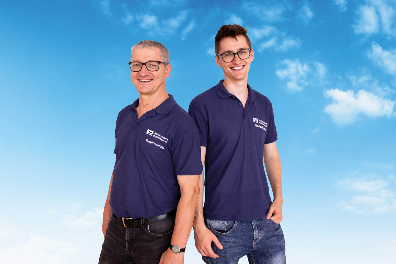Die Kollegen Rudi Hummel und Nicolas Hupfer stehen Seite an Seite strahlend vor einem blauen Himmel.