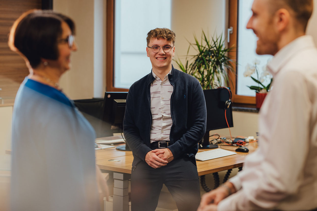Drei Personen tauschen sich im Büro miteinander aus, eine Person lacht in die Kamera