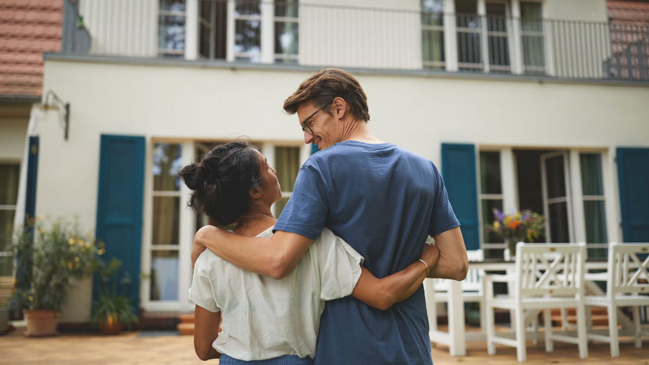 Mann und Frau stehen Arm in Arm vor einem Haus und lächeln sich an