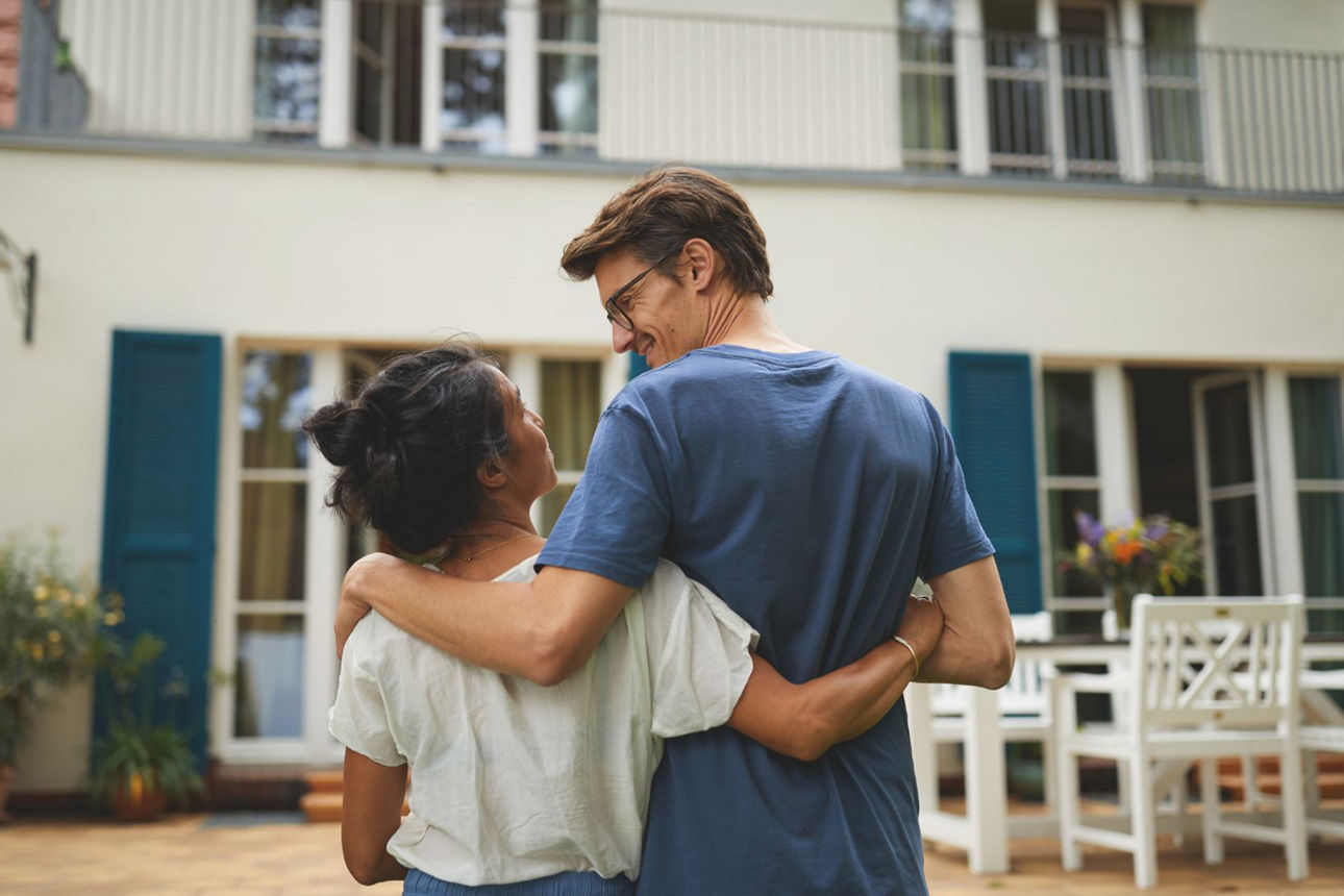 Mann und Frau stehen Arm in Arm vor einem Haus und lächeln sich an