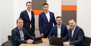 Unser e-Business-Beraterteam - Drei Berater sitzend am Tisch, zwei stehend im Hintergrund 