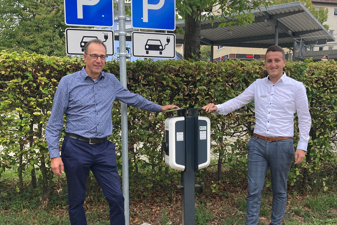Zwei Männer stehen auf dem Parkplatz der Bank neben einer E-Ladestation