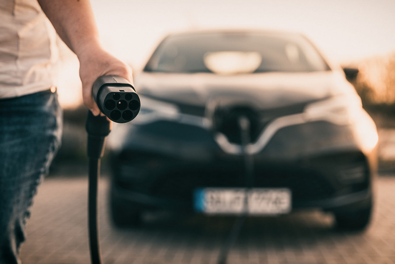 Im Hintergrund sieht man verschwommen ein E-Auto am Ladekabel. Das andere Endstück des Kabels ist im Vordergrund scharf