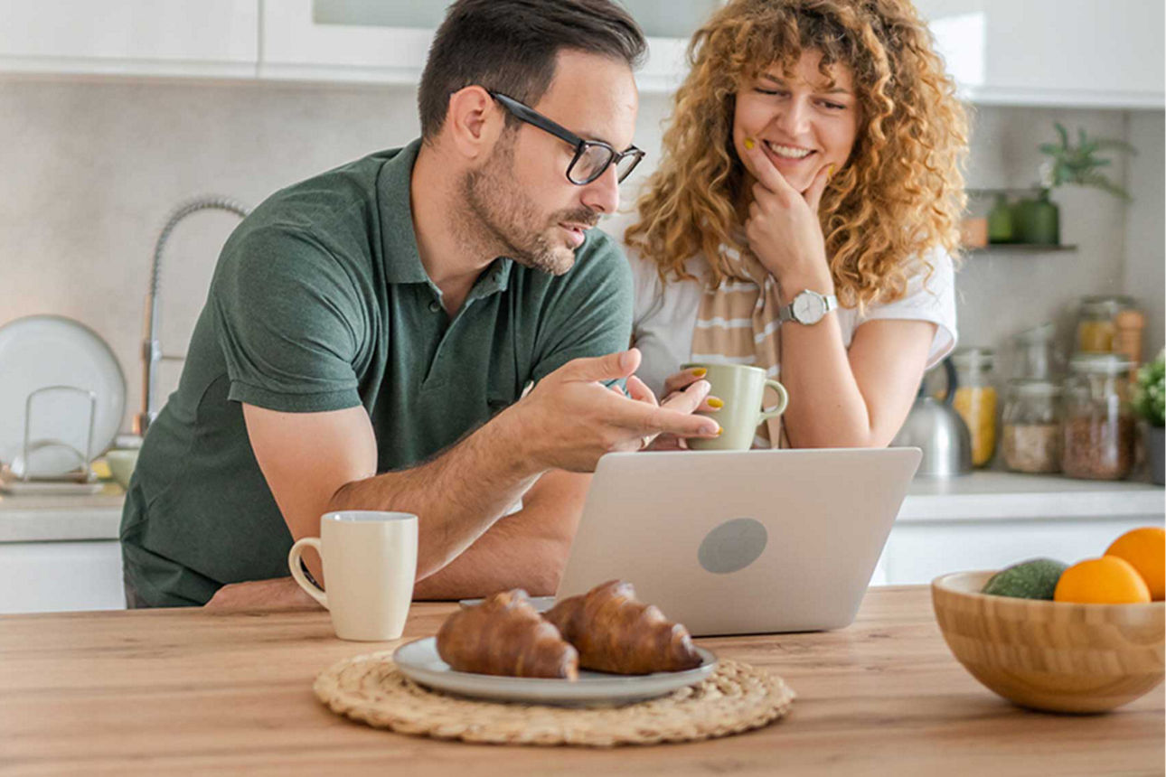 Ein Paar (Mann und Frau) stehen am Küchentisch und schauen gemeinsam auf ein Laptop