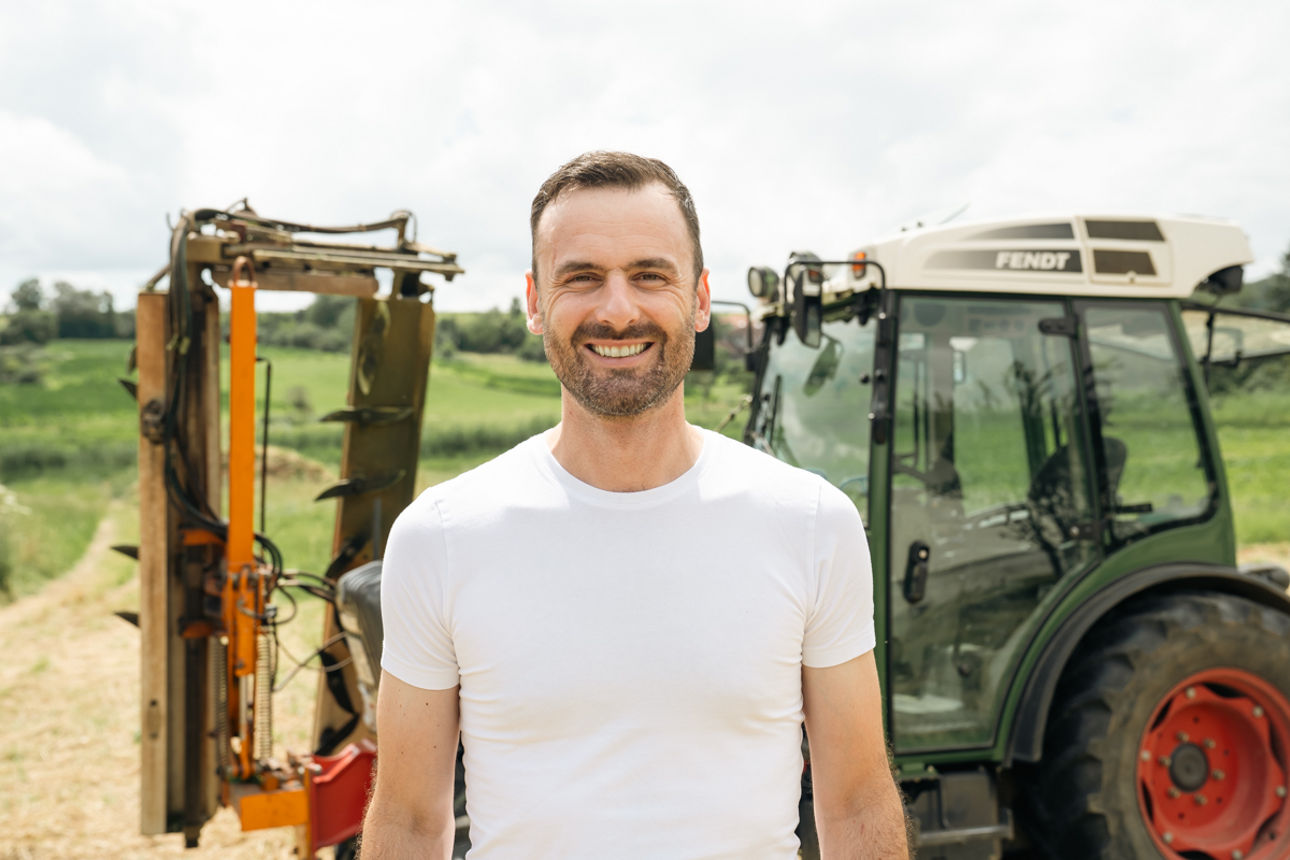 Ein Mann steht auf einem Feld vor einem Traktor, vermutlich in landwirtschaftlicher Umgebung