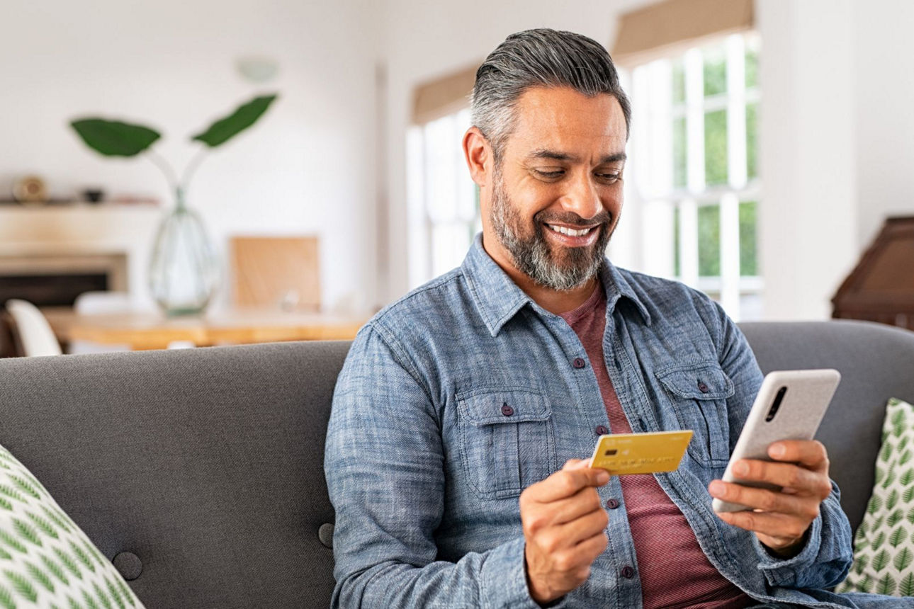 Ein Mann hat eine Bankcard und ein Handy in der Hand und sitzt auf einer Couch