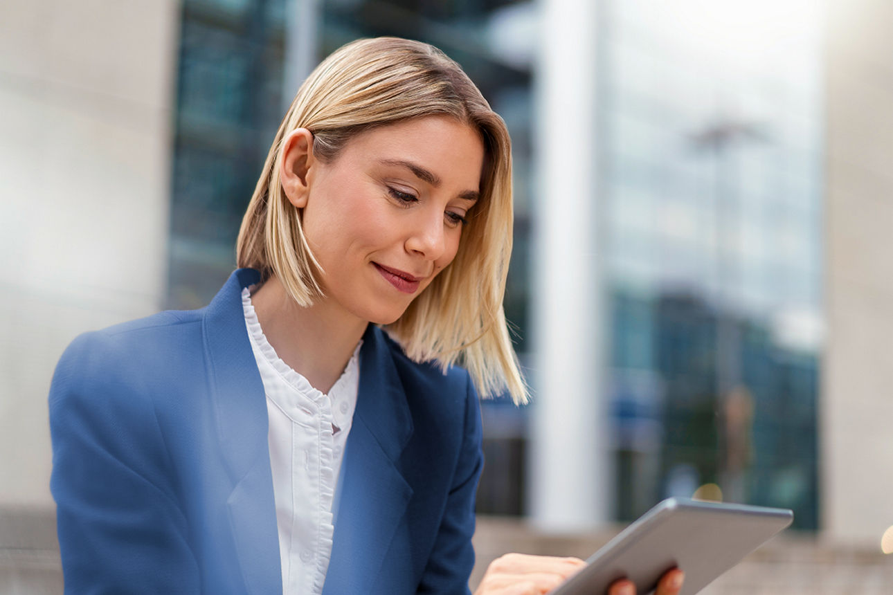 Eine Frau mit blauem Blazer und weißer Bluse tippt auf ein Tablet, das sie in den Händen hält.