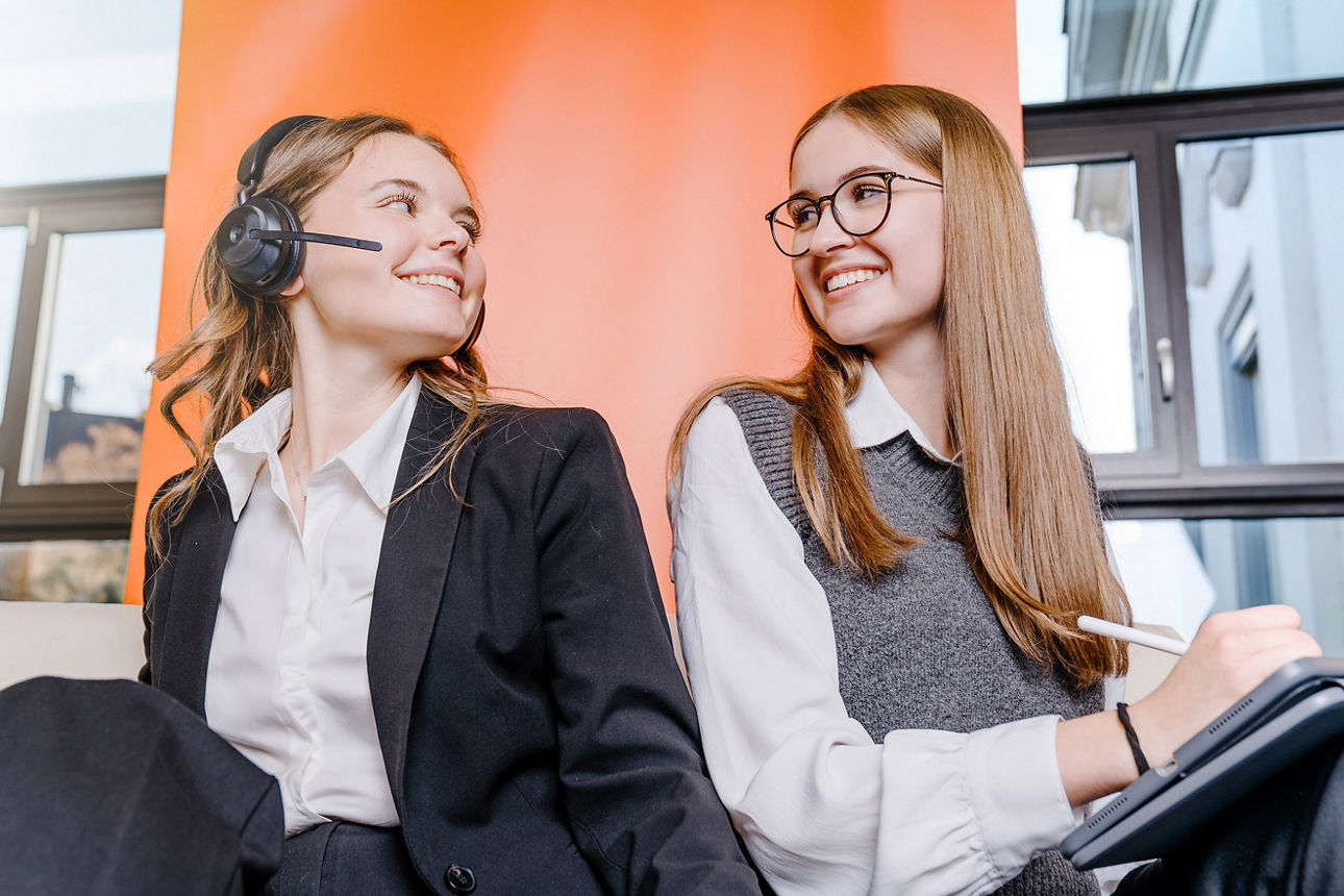 Eine Auszubildende mit Kopfhörer und eine mit iPad lachen sich gegenseitig an