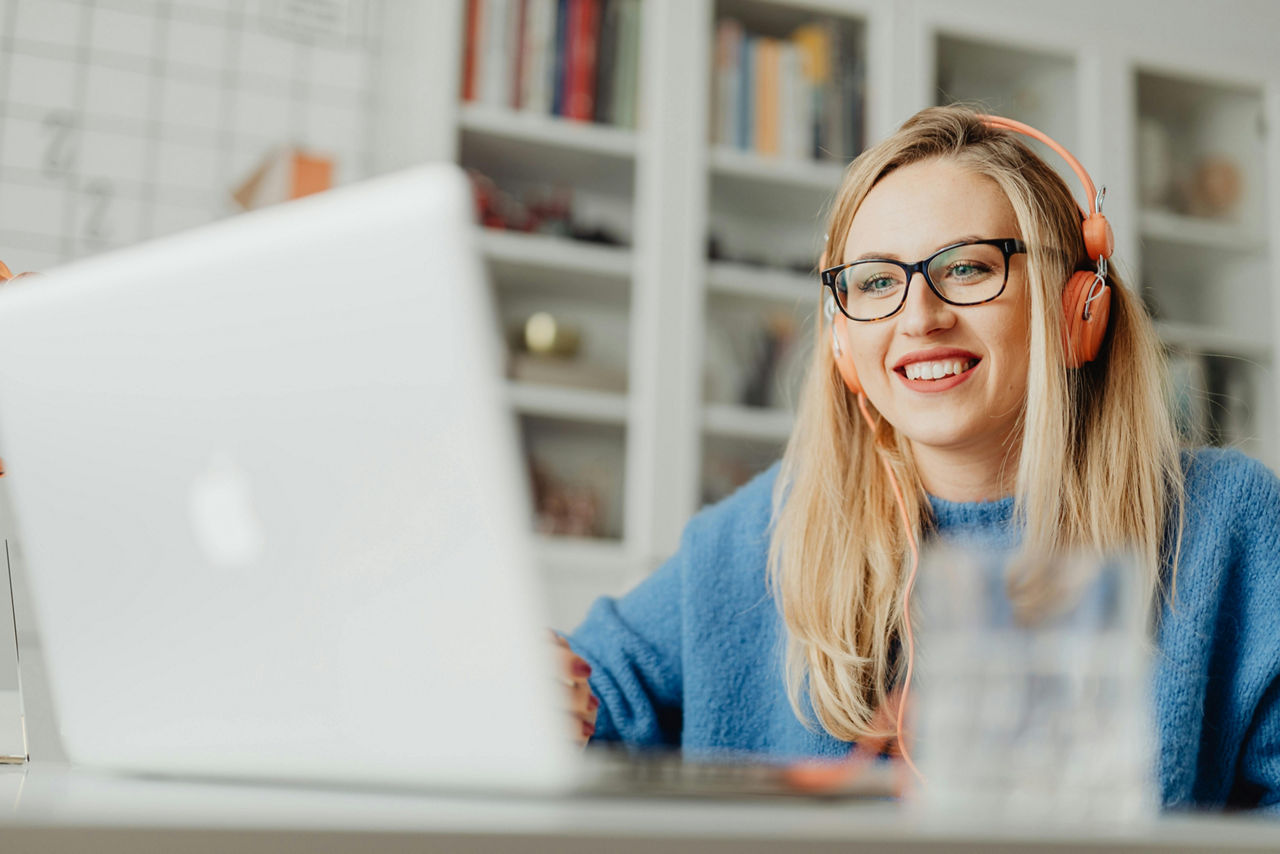 Eine Frau mit Brille und Kopfhörern sitzt lächelnd vor einem Laptop in einem hellen Raum mit Bücherregal im Hintergrund.
