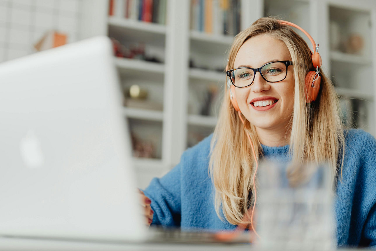 Eine Frau mit Brille und Kopfhörern sitzt lächelnd vor einem Laptop in einem hellen Raum mit Bücherregal im Hintergrund.
