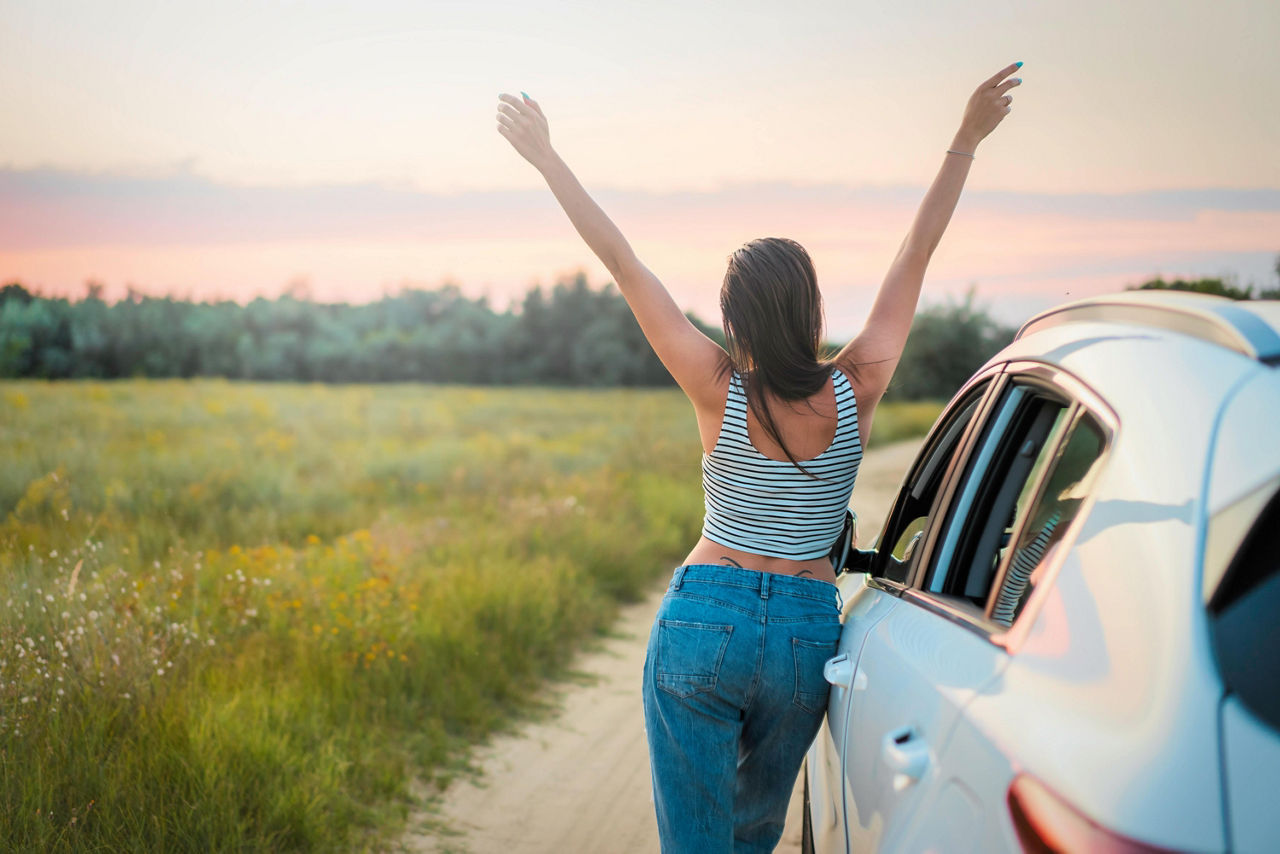 Eine Frau steht neben ihrem Fahrzeug, blick auf eine Wiese und reist vor Freude die Hände hoch