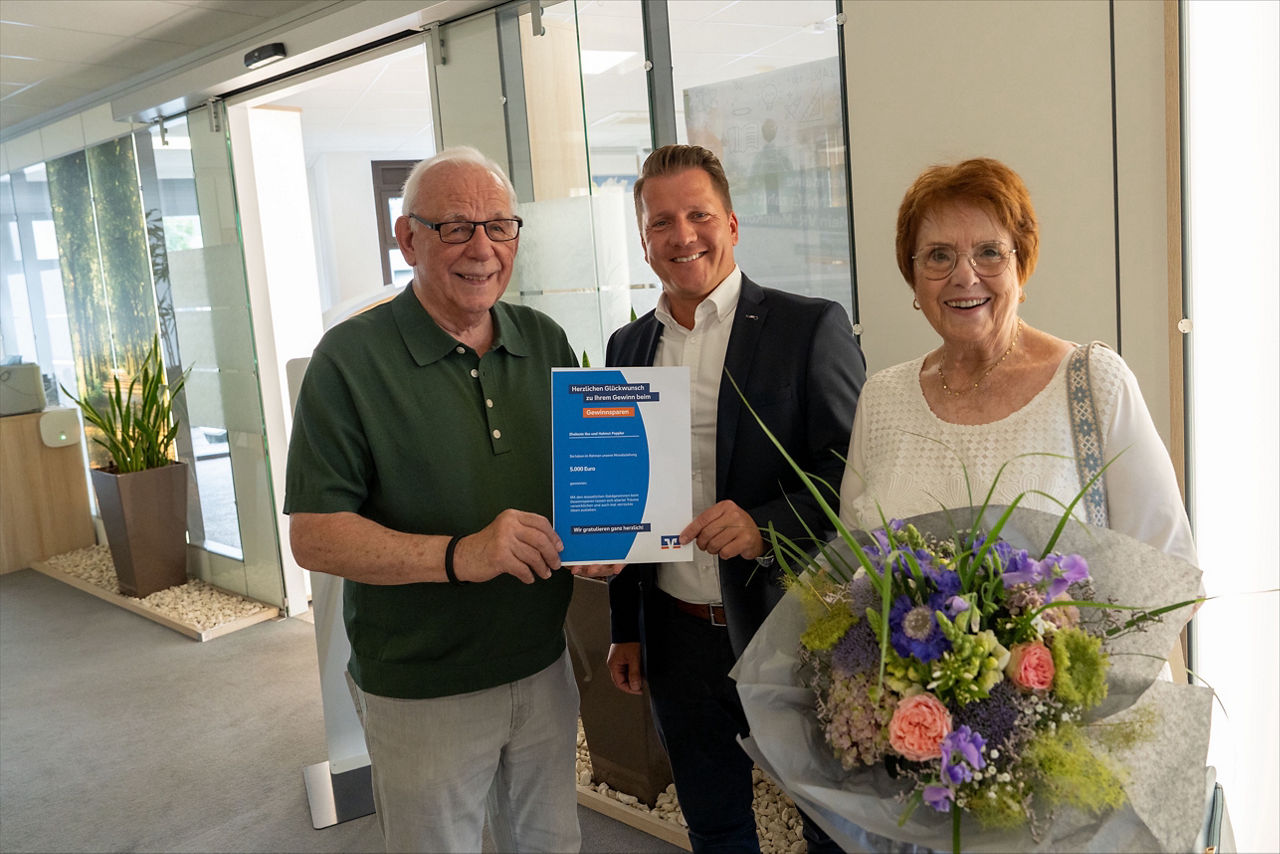 Eine Frau und zwei Männer lachen in die Kamera während sie eine Urkunde und einen Blumenstrauß in den Händen halten