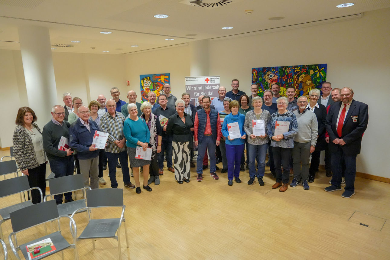 Eine Gruppe von etwa 30 Personen steht in einem hell beleuchteten Raum mit Holzboden und weißen Wänden.