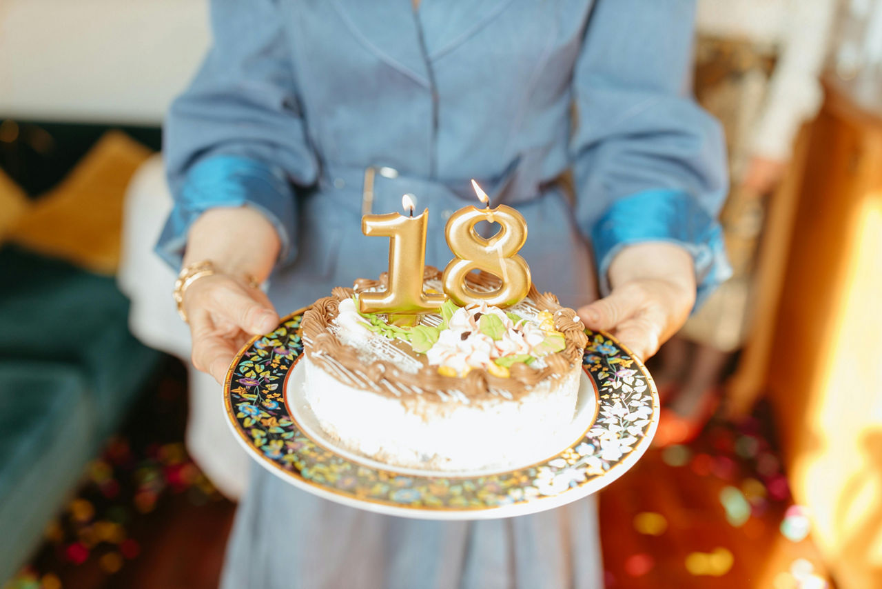Eine Person hält eine Geburtstagstorte mit einer Zahlkerze zum 18. Geburtstag in die Kamera