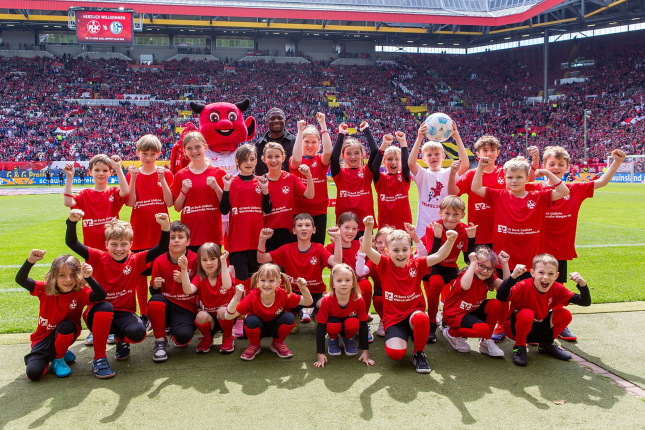 Gruppe von Kindern in roten Trikots posiert am Spielfeldrand eines vollen Stadions für ein Foto.