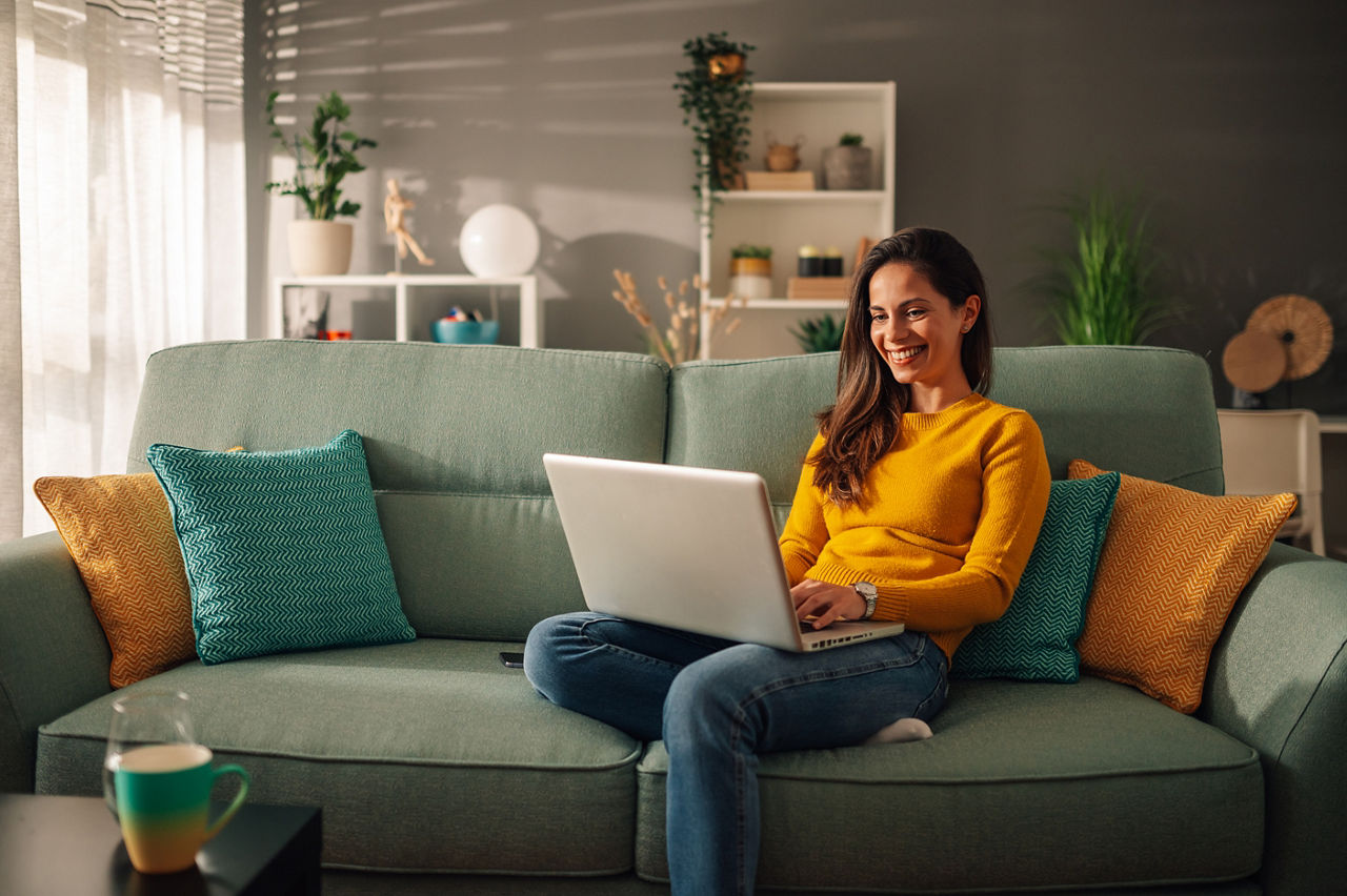 Frau sitzt auf der Couch und schaut lächelnd auf ihren Laptop