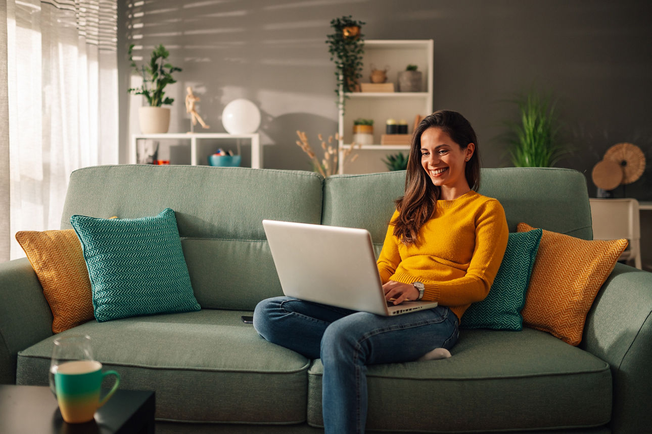 Frau sitzt auf der Couch und schaut lächelnd auf ihren Laptop