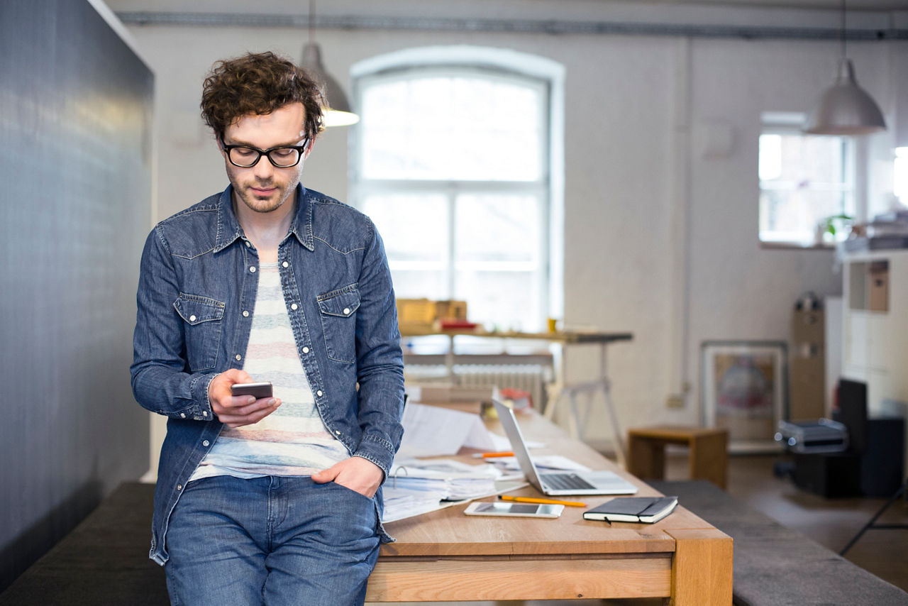 Ein junger Mann hat ein Handy in der Hand und steht vor einem Schreibtisch