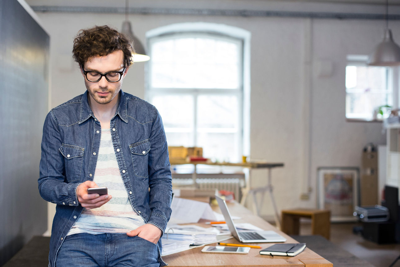 Ein junger Mann hat ein Handy in der Hand und steht vor einem Schreibtisch