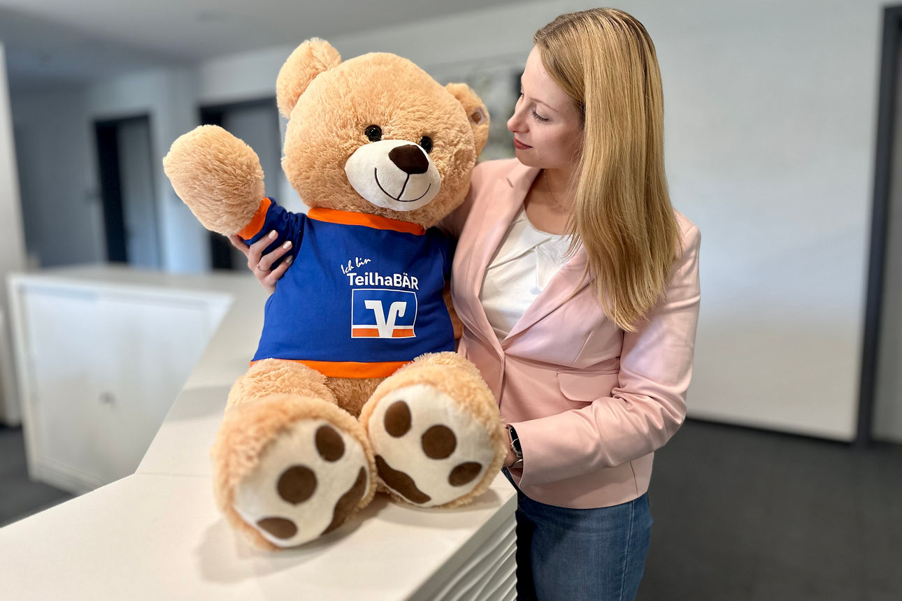 Eine junge Frau im rosafarbenen Blazer hält einen großen Teddybären im Arm, auf dessen T-Shirt steht: Ich bin ein Teilhabär.