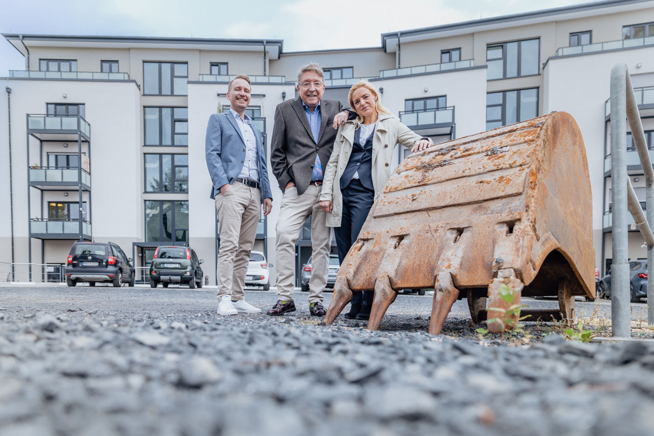 Mehrere Personen stehen im Außenbereich eines Wohnprojekts von ELMLAND1 vor einem fertiggestellten Mehrfamilienhaus. Im Vordergrund ist ein Baumaschinenteil zu sehen, im Hintergrund moderne Wohngebäude mit Balkonen, Stellplätzen und klarer Fassadengestaltung.
