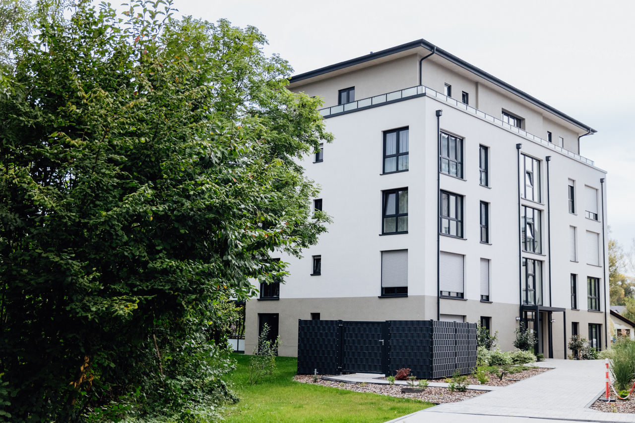 Außenansicht eines mehrgeschossigen Wohngebäudes des Projekts ELMLAND1, eingebettet in begrünte Außenflächen mit Wegen und Bepflanzung. Die Aufnahme zeigt die moderne Fassadengestaltung und die Einbindung des Gebäudes in die naturnahe Umgebung des Wohnensembles.