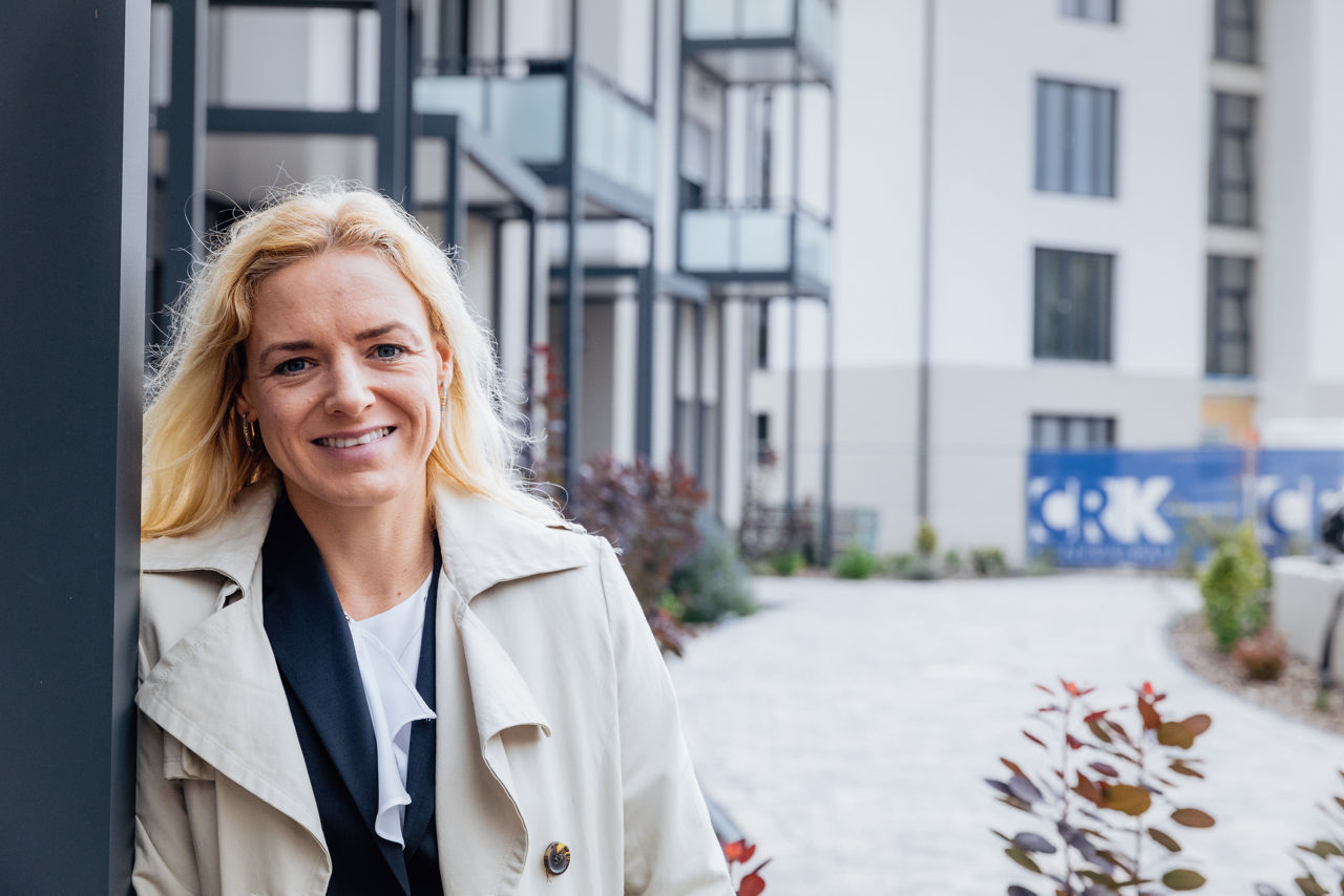 Eine Person steht im Außenbereich eines Wohngebäudes des Projekts ELMLAND1. Im Hintergrund sind Balkonstrukturen, Fensterflächen und Wegeführung sichtbar, die den hochwertigen Ausbau und die moderne Architektur des Wohnensembles zeigen.