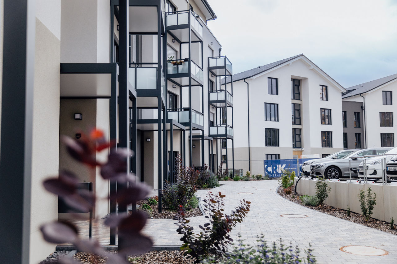 Blick entlang eines gepflasterten Weges durch den Außenbereich des Wohnprojekts ELMLAND1. Die Aufnahme zeigt mehrgeschossige Wohngebäude mit Balkonen, gestaltete Grünflächen sowie Stellplatz‑ und Erschließungsbereiche als Teil des fertigen Wohnensembles.