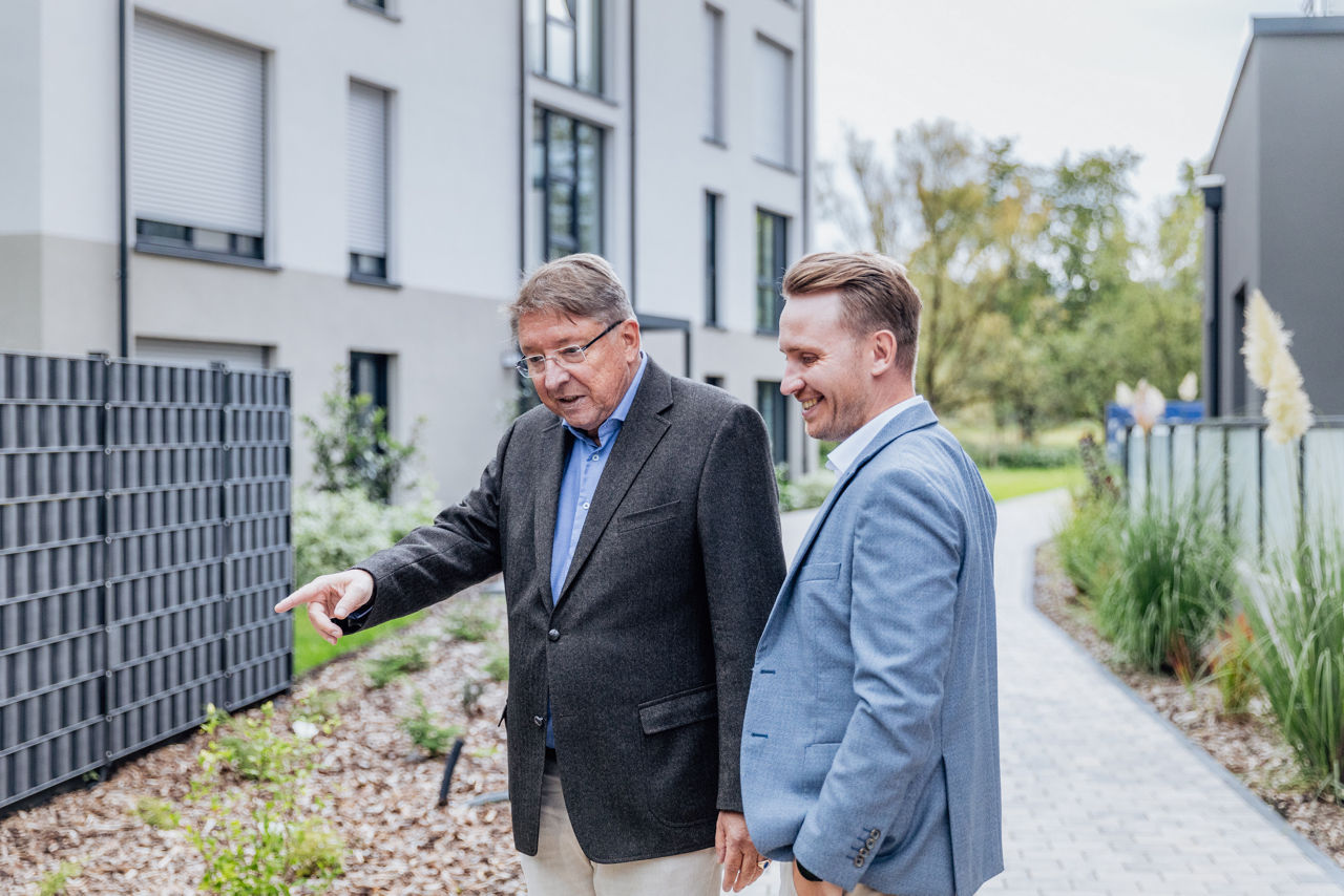 Zwei Personen stehen im Außenbereich eines Wohnprojekts von ELMLAND1 zwischen fertiggestellten Wohngebäuden und gestalteten Grünflächen. Die Aufnahme zeigt eine Projektsituation im bereits realisierten Wohnumfeld mit moderner Architektur und klarer Wegeführung.
