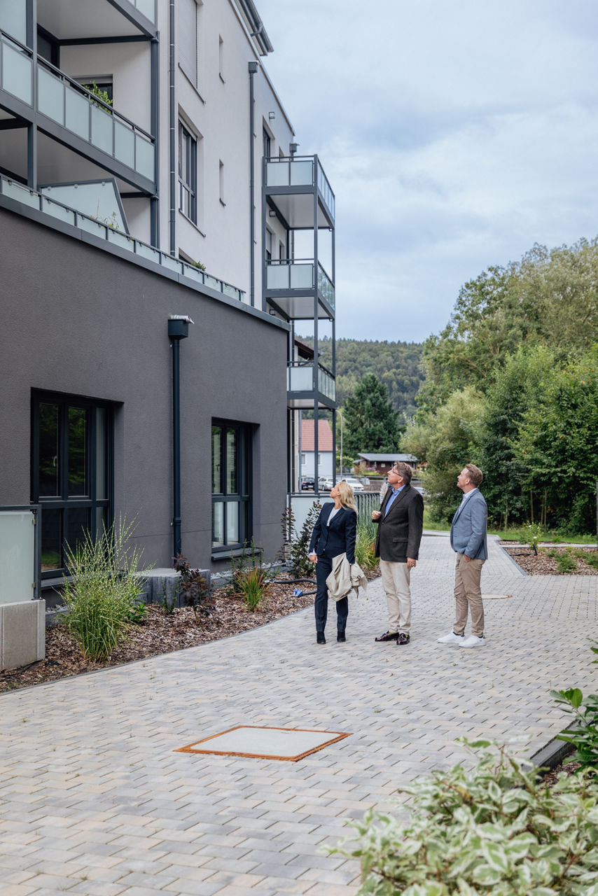 Mehrere Personen stehen im Außenbereich eines Wohngebäudes des Projekts ELMLAND1 und betrachten die Fassade sowie die Balkonstrukturen. Die Aufnahme zeigt den fertiggestellten Gebäudekomplex mit gepflasterten Wegen, Grünflächen und moderner Architektur im Wohnumfeld.