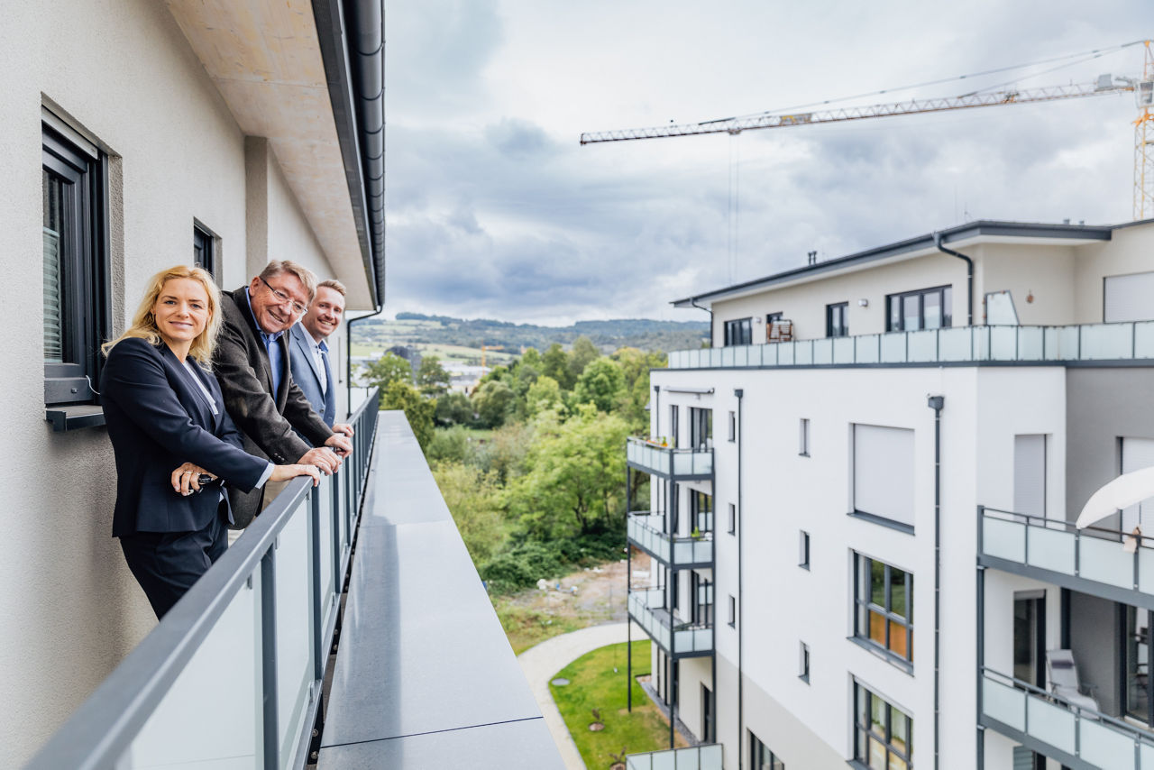 Mehrere Personen stehen auf einem Balkon innerhalb des Wohnprojekts ELMLAND1 und blicken auf gegenüberliegende Wohngebäude sowie die umgebende Landschaft. Die Aufnahme zeigt die Architektur, die Höhenstaffelung der Gebäude und die räumliche Einbindung des Projekts in das Umfeld.