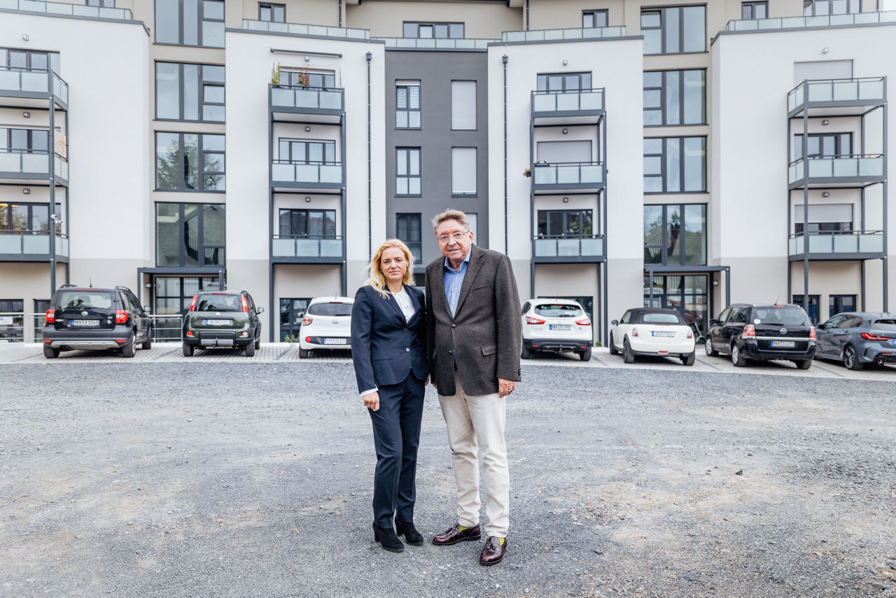 Zwei Personen stehen im Vordergrund eines fertiggestellten Wohngebäudes des Projekts ELMLAND1. Im Hintergrund sind mehrgeschossige Wohnhäuser mit Balkonen, Fensterflächen und Stellplätzen zu sehen, die die moderne Architektur und den abgeschlossenen Bauzustand des Wohnensembles zeigen.
