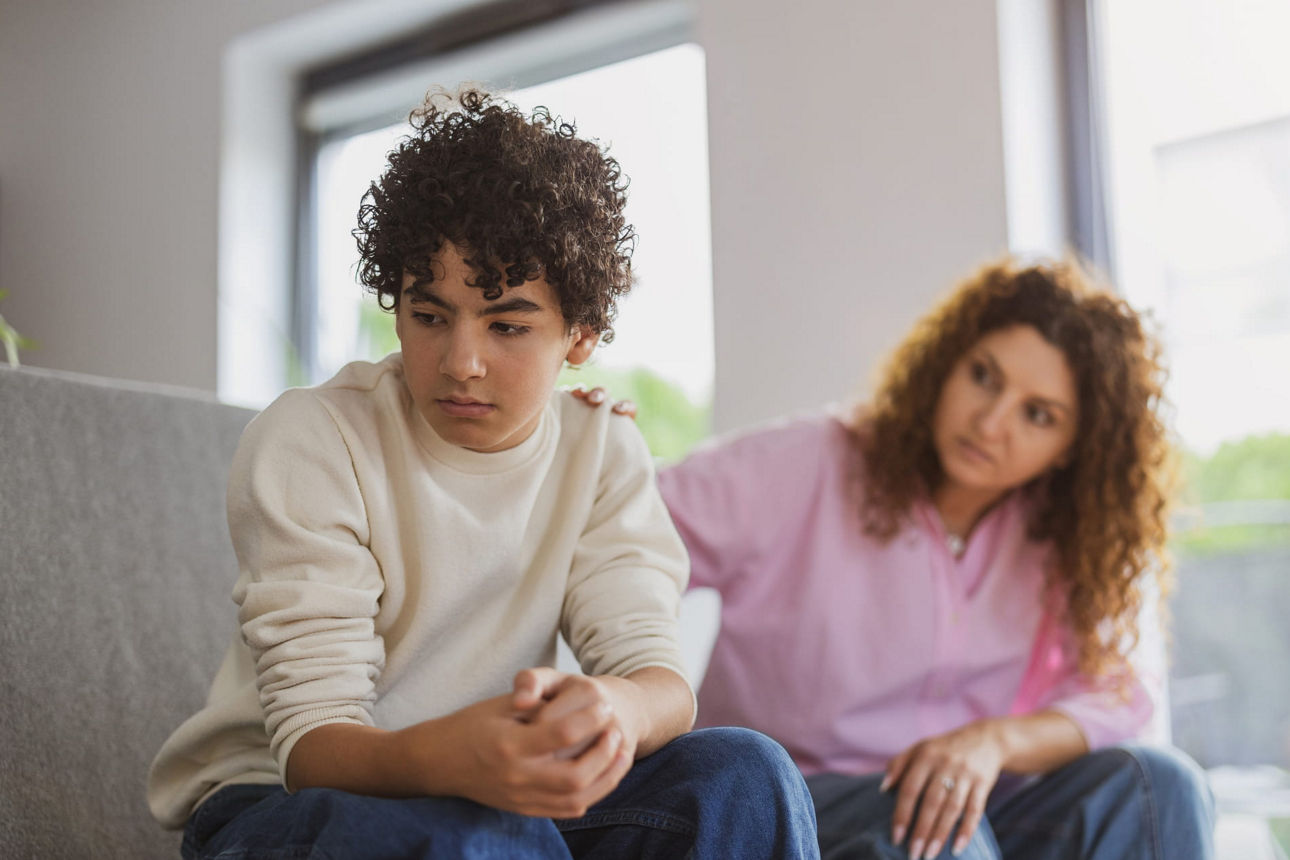 Ein frustierter Jugendlicher sitzt auf dem Sofa, seine Mutter ist im Hintergrund zu sehen.