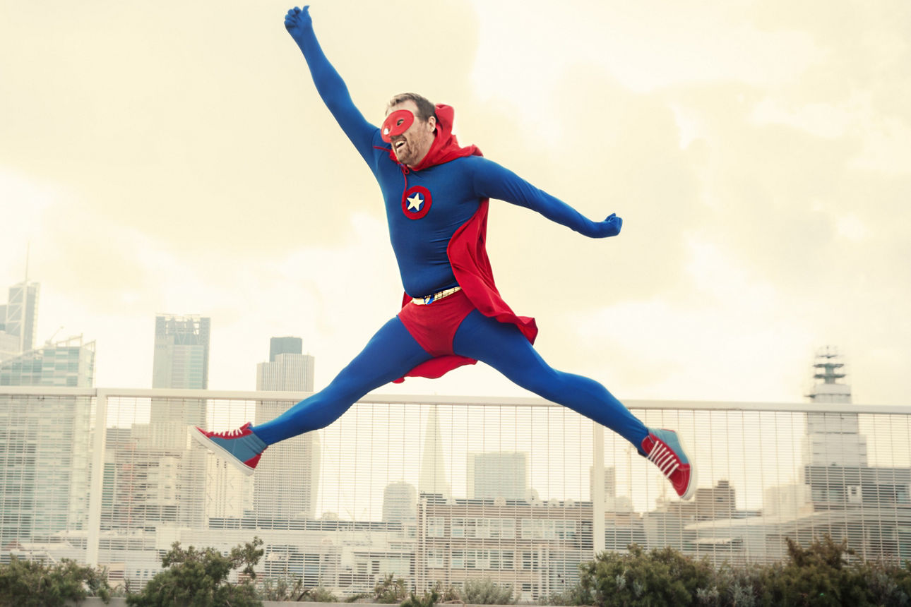Mann im Supermann-Kostüm springt auf einem Dach. Im Hintergrund die Skyline einer Stadt.