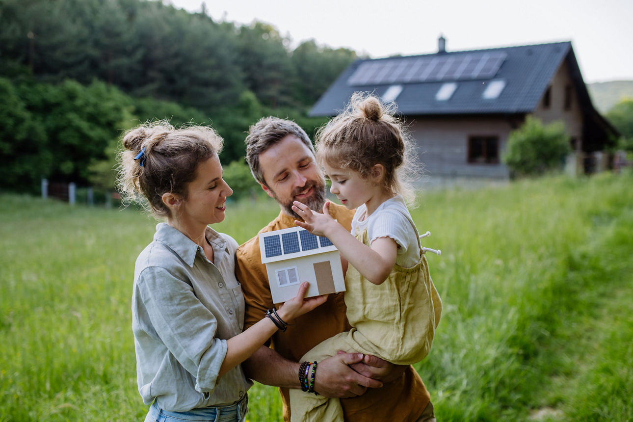 Dreiköpfige Familie steht auf der Wiese vor einem Einfamilienhaus mit Solarpanelen
