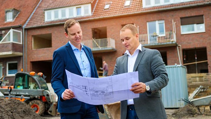 Zwei Energieberater der Bank stehen vor einem Haus, welches saniert wird. In der Hand halten Sie den Grundriss.