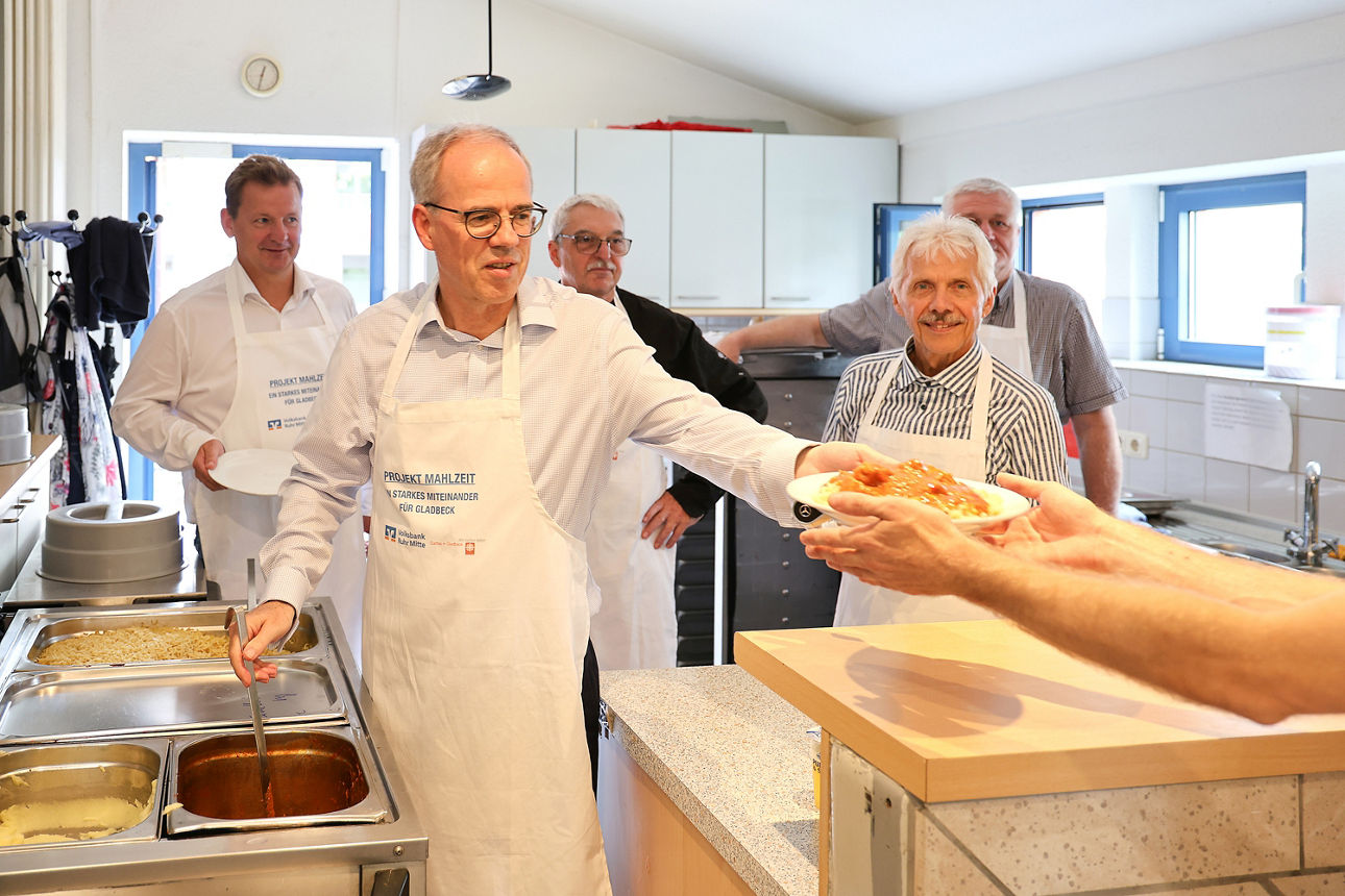 Zu sehen sind 5 Personen, die in der offenen Küche eines Gemeindezentrums stehen. Alle tragen eine weiße Küchenschütze mit dem Aufdruck "Projekt Mahlzeit". Im Vordergrund steht unser Vorstand Herr Abrahams und überreicht gerade einen vollen Teller mit Essen an eine Person außerhalb des Bildes. 