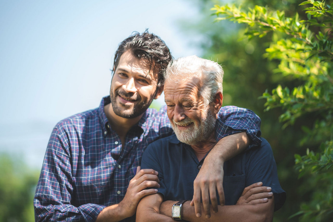 Erwachsener Sohn legt seinen linken Arm um seinen älteren Vater. Beide lächeln zufrieden. Im Hintergrund ist ein Busch.