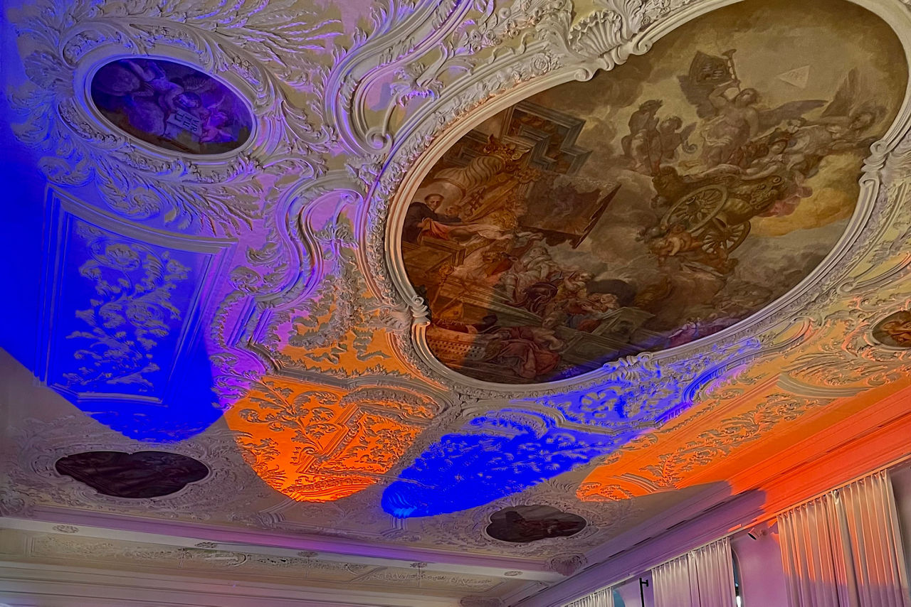 Fote der reich verzierten Stuckdecke mit großem Gemälde in der Mitte; angestrahlt mit orangenen und blauen Scheinwerfern