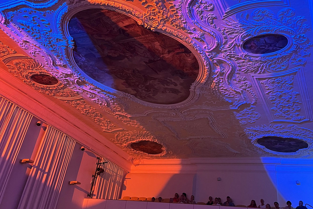 Ausschnittsfoto der Stuckdecke des Freisinger Asamsaals eingetauch in orangenes und blaues Licht, den Farben der Volks- und Raiffeisnebanken