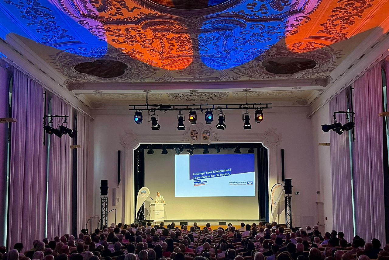 von hinten auf die Bühne fotografiert der vollbesetzte Asamsaal in Freising; auf der Bühne steht links ein kleines Rednerpult mit einem Mann dahinter, an die Wand wird ein Ausschnitt einer Präsentation projiziert; die Stuckdecke wird orange-blau beleuchtet
