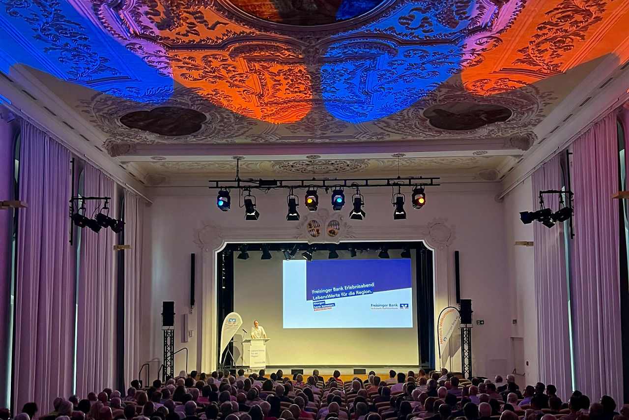 Blick vom hinteren Teil des Saals auf die Bühne. Im Vordergrund Publikum im Dunklen, auf hellerleuchteter Bühne steht ein Redner mit Pult, die Decke wird mit blauen und orangenen Scheinwerfern angestrahlt