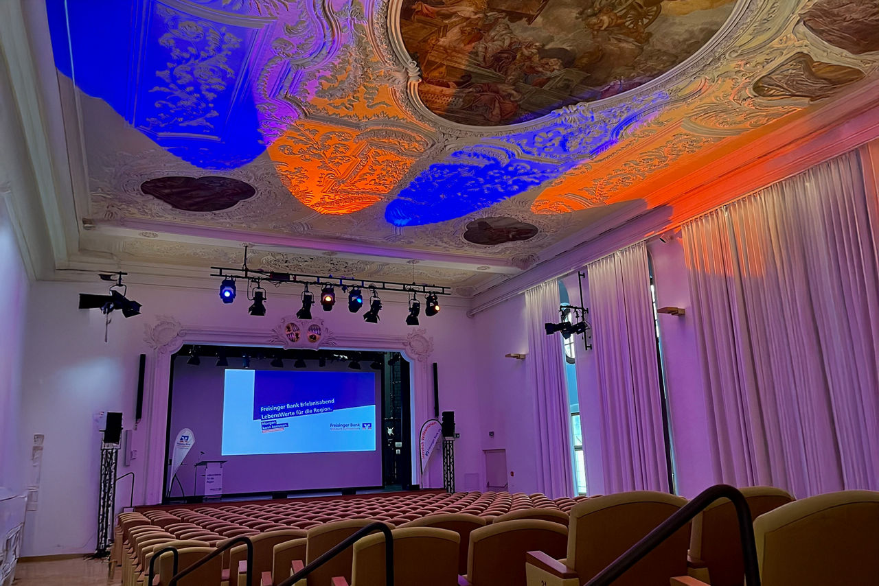 Asamsaal Freising in Vorbereitung auf den Erlebnisabend der Freisinger Bank; im Vordergrund leere Sitzreihen von hinten mit Blick auf die Bühne; die Stuckdecke ist mit  orangenen und blauen Scheinwerfern angestrahlt