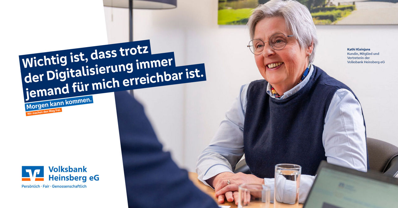 Eine Frau sitzt lächelnd im Büro, im Vordergrund sitz ein Mann im blauen Anzug.