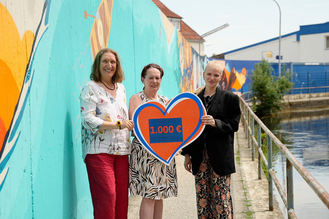 Drei Frauen stehen bei bunt bemalten Mauer und halten ein Spendenherz über 1000 Euro. Rechts ein Geländer und Gewässer