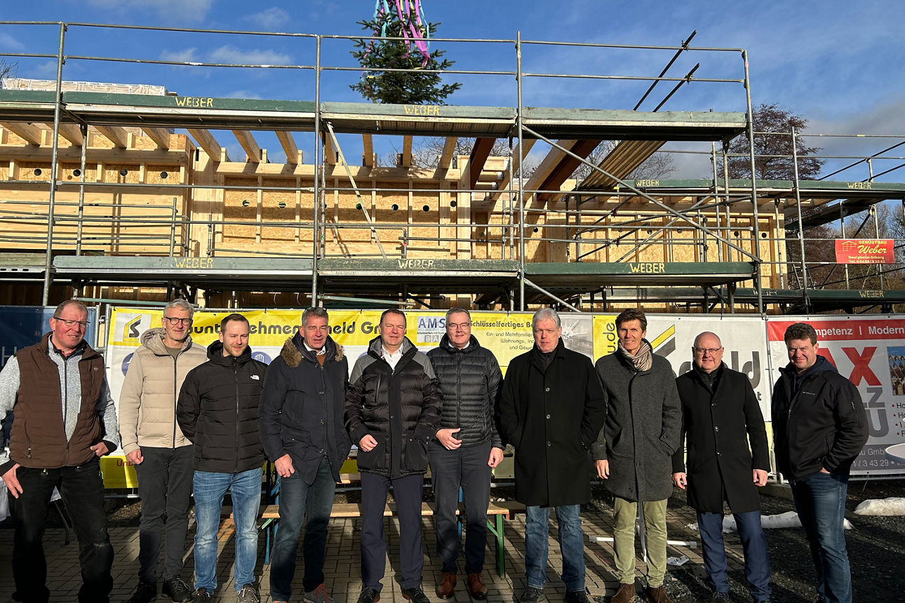 Gruppe von elf Männern in Winterjacken steht vor einem Rohbau mit Gerüst und Werbebannern.