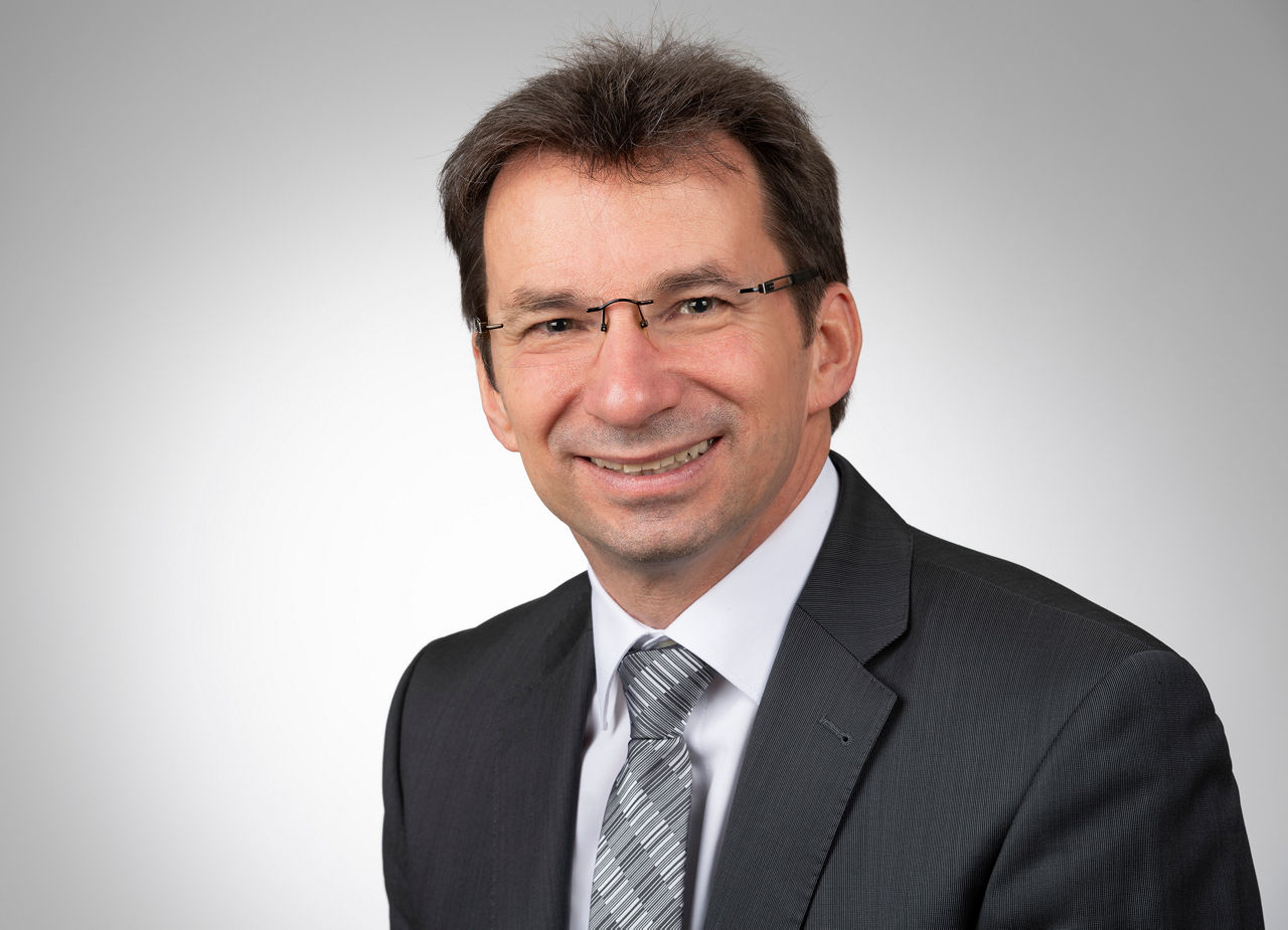 Mitarbeiter mit kurzen dunklen Haaren und Brille mit Anzug lächelt in die Kamera.