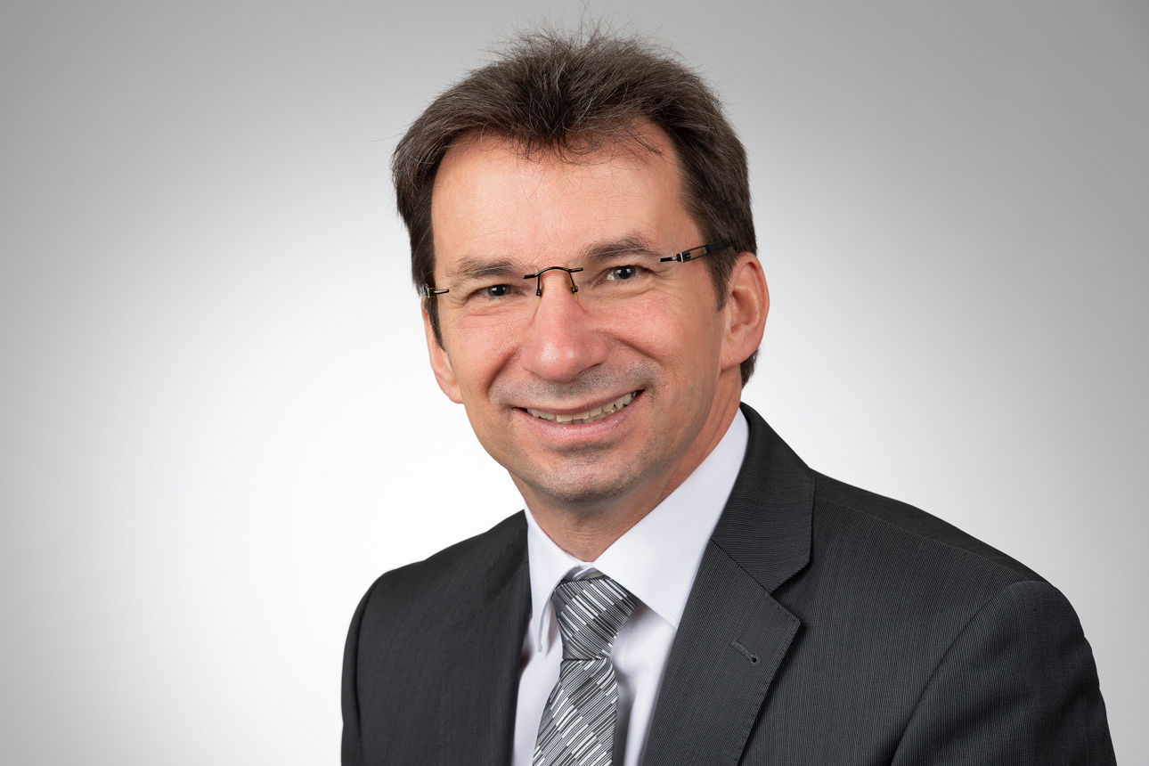 Mitarbeiter mit kurzen dunklen Haaren und Brille mit Anzug lächelt in die Kamera.